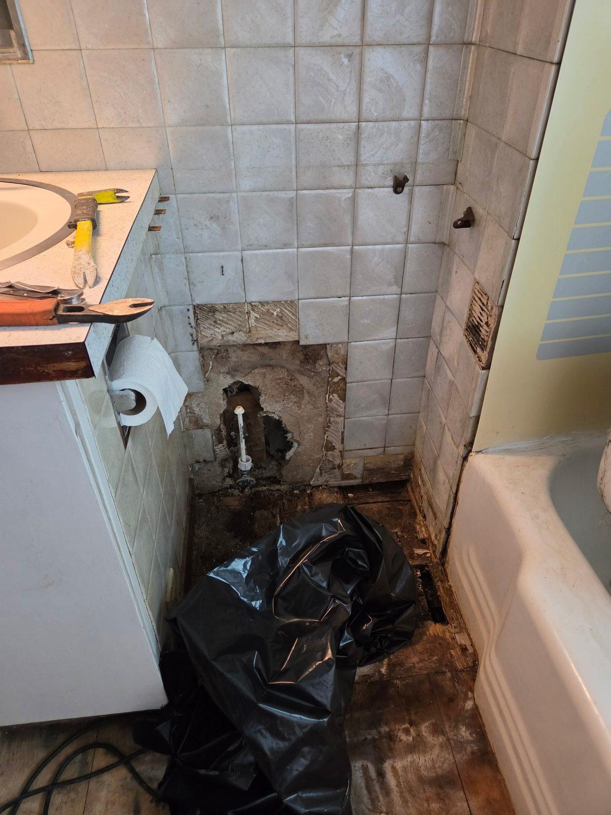 Bathroom with wall tile removed, showing rotten subfloor and exposed pipework beside the tub