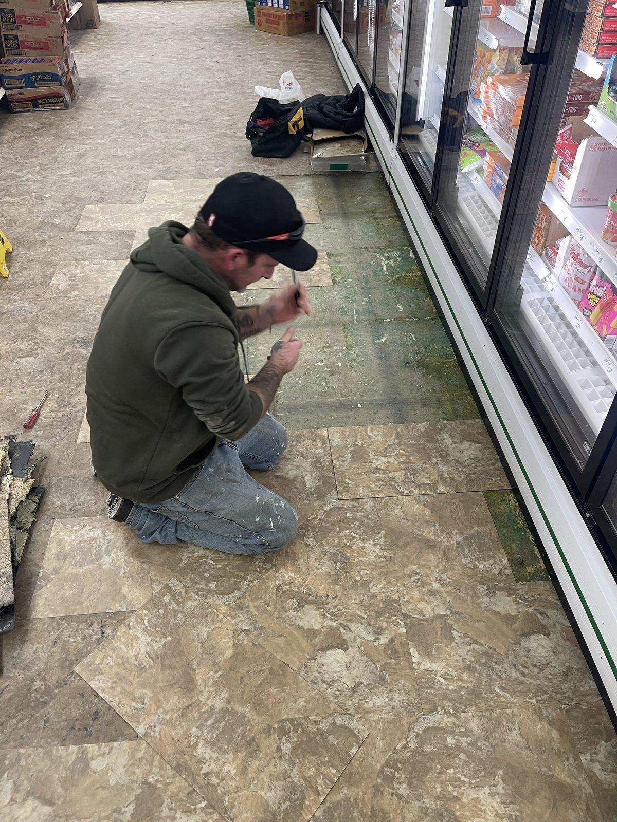 Tradesman laying stone-effect floor tile in a shop aisle beside the chiller cabinets